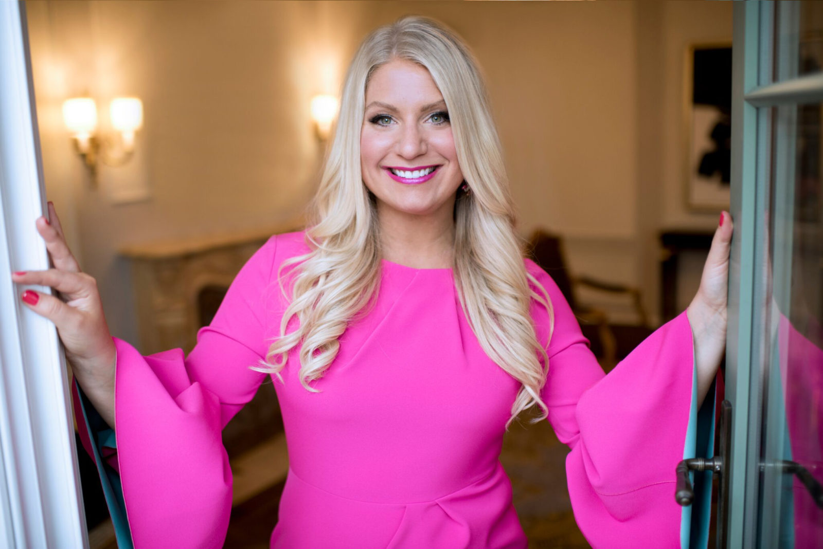 Emily Williams in a pink dress, smiling as she opens a pair of french doors