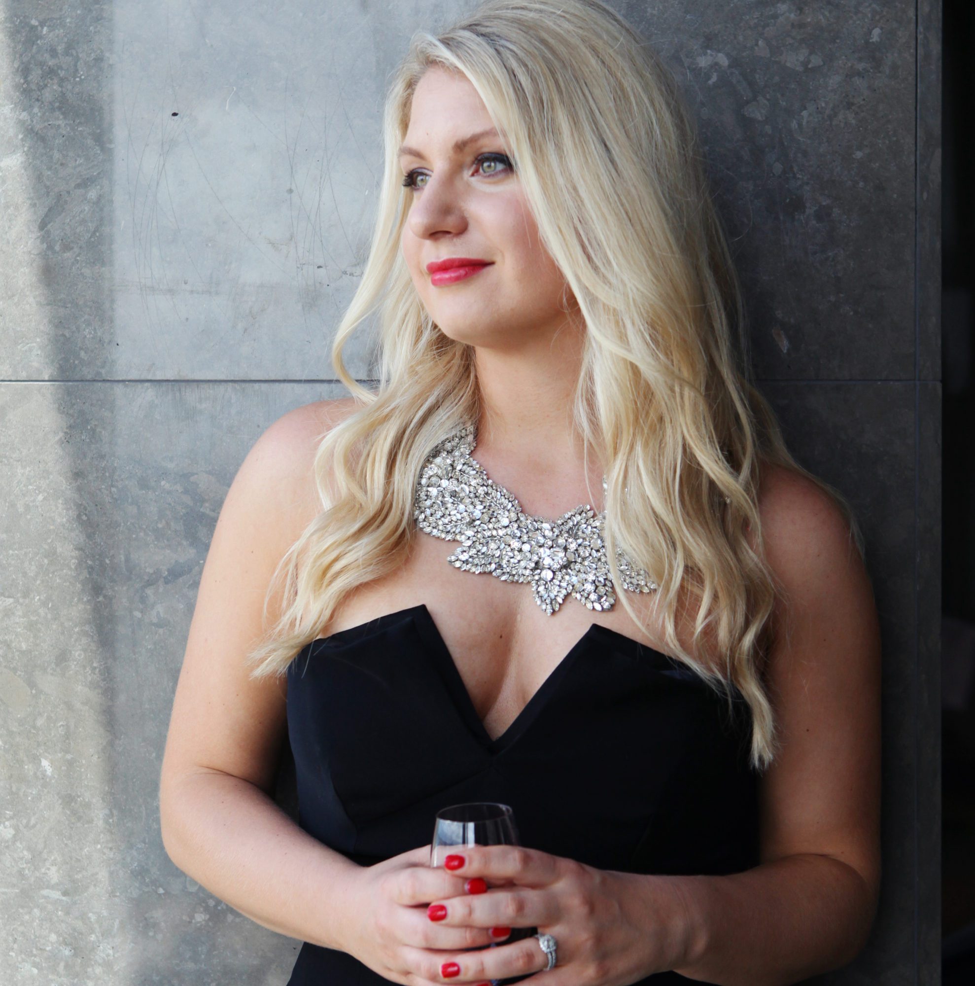 Emily Williams Holding a Champagne Glass while wearing a black dress and silver necklace
